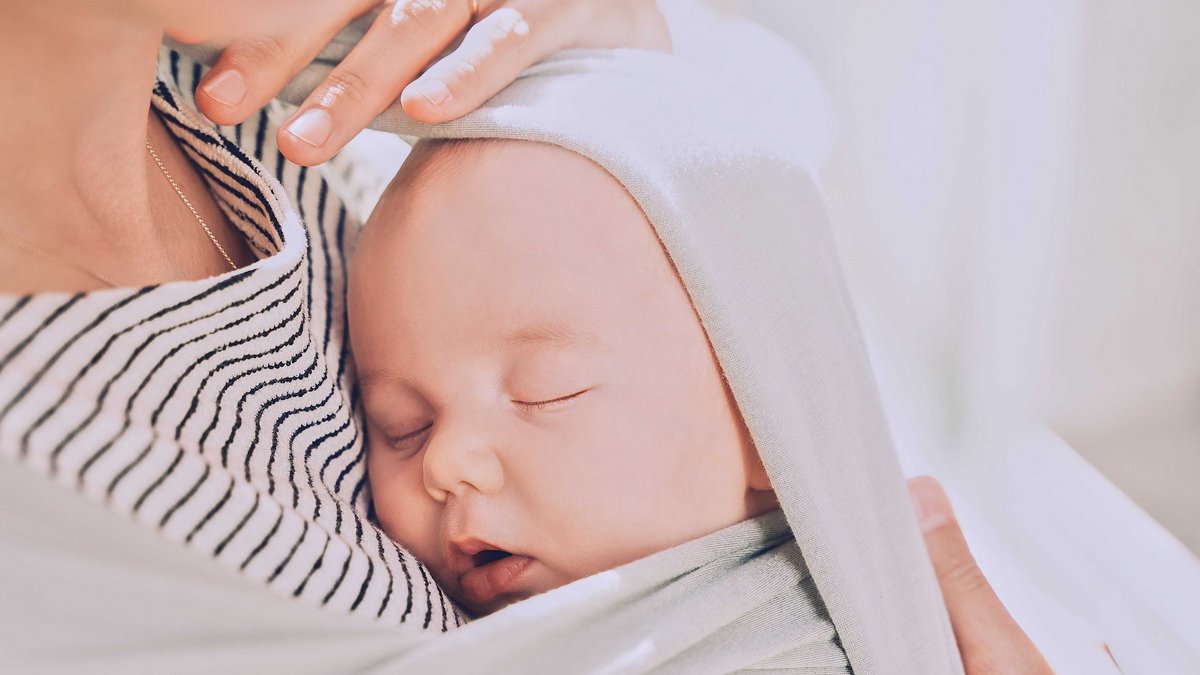 Ein Baby liegt in ein Tuch gewickelt an der Brust einer Person und schläft.
