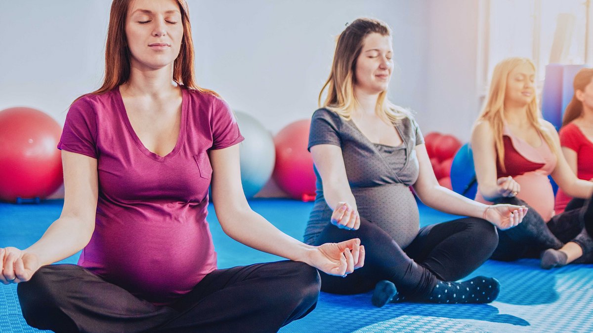 Eine Reihe an schwangeren Frauen sitzen im Schneidersitz auf dem Boden und meditieren mit geschlossenen Augen.