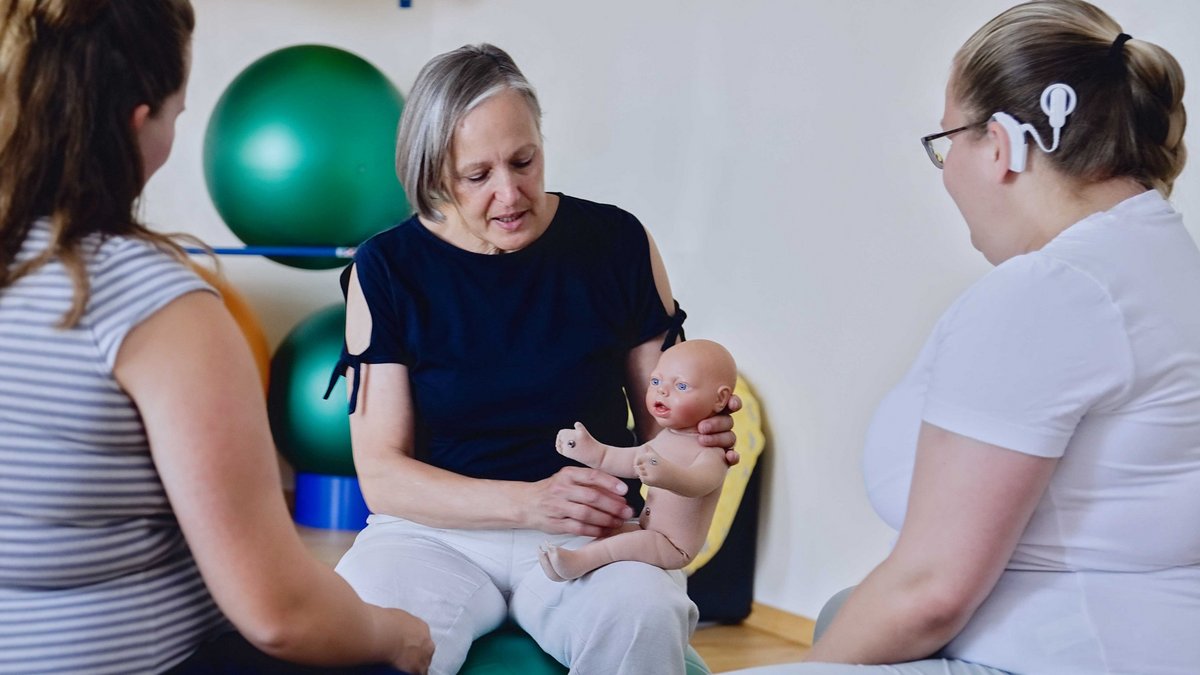 Eine Frau sitzt mit einer Baby-Puppe auf dem Bein und erklärt etwas, zwei weitere Frauen sitzen vor ihr und schauen zu.