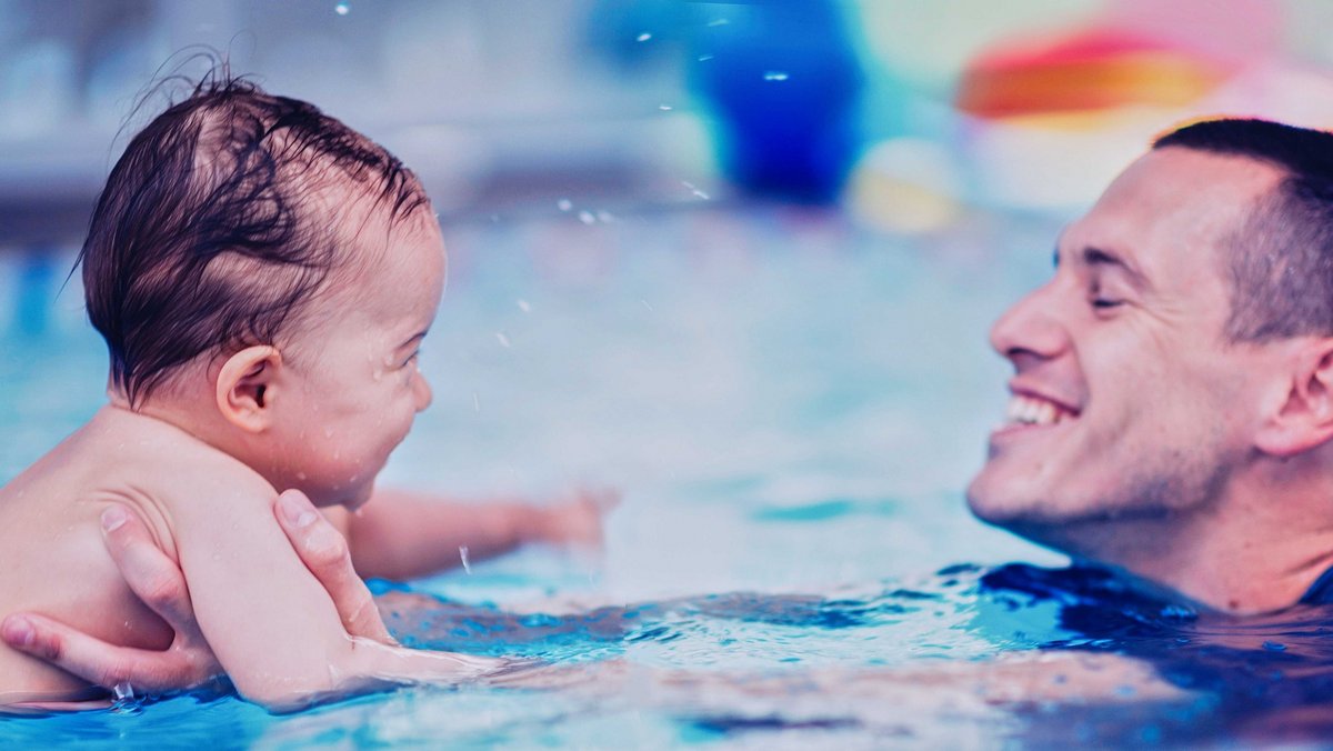 Ein Mann schwimmt im Wasser und blickt lächelnd auf ein Baby, welches er vor sich über Wasser hält.