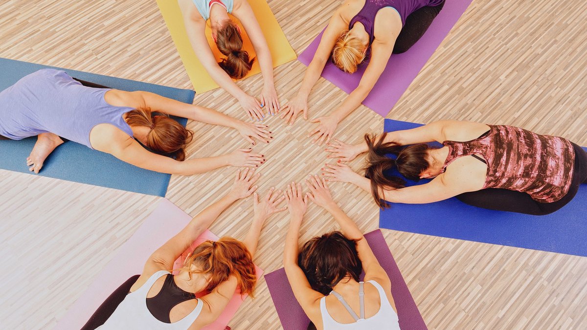 Sechs Frauen liegen im Kreis mit gesenktem Kopf auf dem Bauch auf Yogamatten und bilden mit ihren Händen in der Mitte einen Kreis.