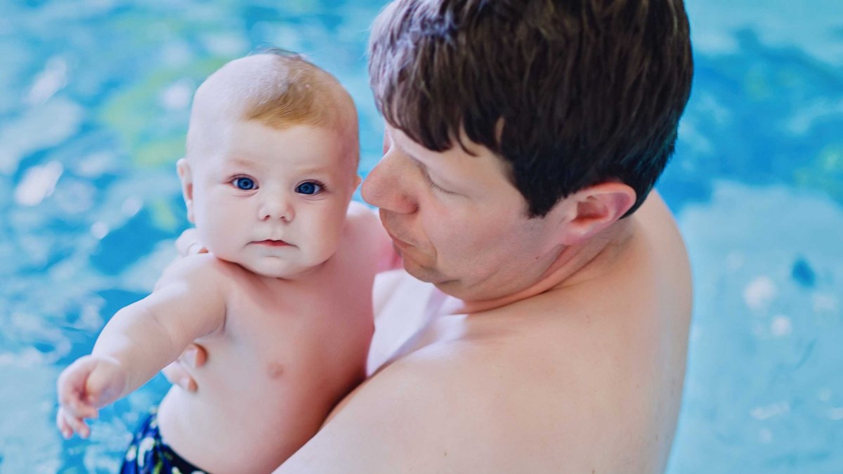Ein Vater steht mit seinem Baby auf dem Arm im Wasser. Das Baby blickt in die Kamera.
